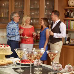 4 people chatting, holding cocktails and food, in front of a home bar, with platters of food in front